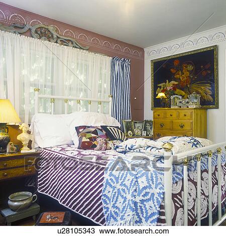 Stock Image - BEDROOM - White brass bed. Stencil border on wall, one wall is terracotta and another is cream, large oil painting, bed stacked with vintage chenille bedspreads, crazy quilt pillows, striped curtains, sheer white curtains, eclectic mix, birds eye mapl. Fotosearch