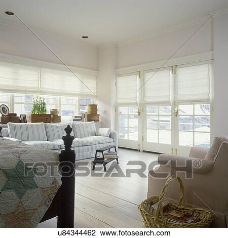 Stock Image - BEDROOMS: Light and Airy bedroom, view towards windows with white fabric shades. Wood floor. Sofa seating area, partial view of bed on left, casual fresh country style. Fotosearch