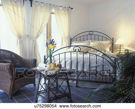 BEDROOMS: Natural cotton fabrics, iron bed set at angle, tab top curtains, natural wicker club chair on left, bamboo table set with breakfast, flowers, tile floor, Green design View Large Photo Image Picture - BEDROOMS: Natural cotton fabrics, iron bed set at angle, tab top curtains, natural wicker club chair on left, bamboo table set with breakfast, flowers, tile floor, Green design. Fotosearch