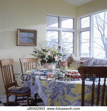 Stock Image - BREAKFAST AREA: Set for dessert, oak farm chairs, vintage linens, floral arrangement, soft pastels. Fotosearch