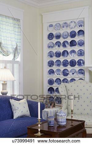 Stock Image - Collection Display: Corner of room with cr? me wingback chair & blue sofa in front of built in shelving unit displaying blue and white Bing & Grondahl Christmas plates. Fotosearch