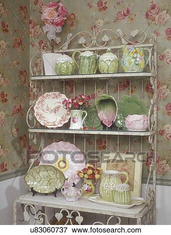 Stock Photo - COLLECTION DISPLAYS: A distressed white metal bakers rack serves as a display for a collection of majolica and garage sale china. Rose patterned wall paper. . Fotosearch