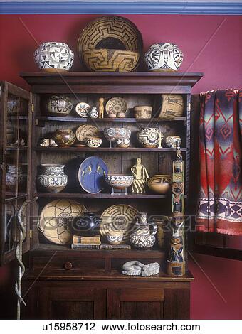 Stock Image - COLLECTION DISPLAYS - Antique cupboard filled with Native American pottery and artifacts, Acoma Ollas early 1900's, walking stick with snake, blanket, red painted walls.. Fotosearch