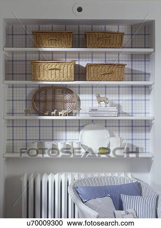 Stock Image - COLLECTION DISPLAYS: Detail of recessed wall shelves with natural wicker baskets, collection of vintage toy sheep, white radiator, white wicker chair, blue and white large check wallpaper in recessed area, set of white small cups. Fotosearch