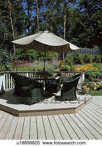 Stock Image - Decks: Dark green wicker furniture sits around a umbrella table on raised platform on deck area, railing, flower garden in bloom in background. . Fotosearch