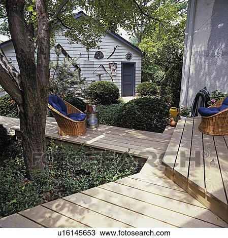 Stock Image - DECKS: Deck Walkway Through Landscaped back yard, wicker basket chairs w/ blue cushions, wall of garage with display of antique farm implements.. Fotosearch
