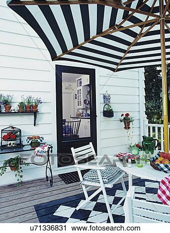 Stock Image - DECKS: White patio table and chairs. Black and white checker board design painted under eating area, potted plants, kitchen in view thru open door.. Fotosearch