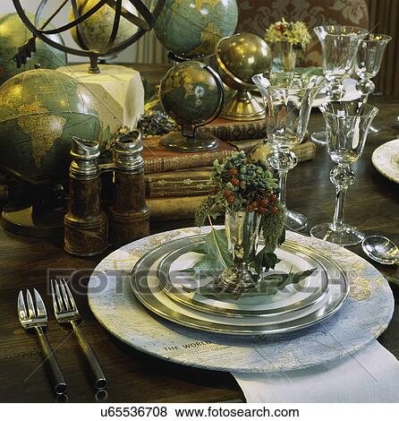 Stock Photo - Detail of a place setting, travel theme with globes, white plates edged with pewter, leather books, maps, vintage binoculars fall or winter season. Fotosearch