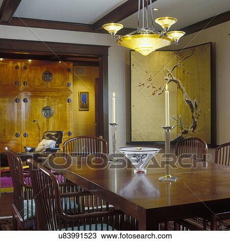 Stock Image - DINING ROOM - Decor Oriental motif. Prairie style architecture, Deco chandelier, gold folding screen, rectangular table, large Oriental cabinet with Eames chair in background, . Fotosearch