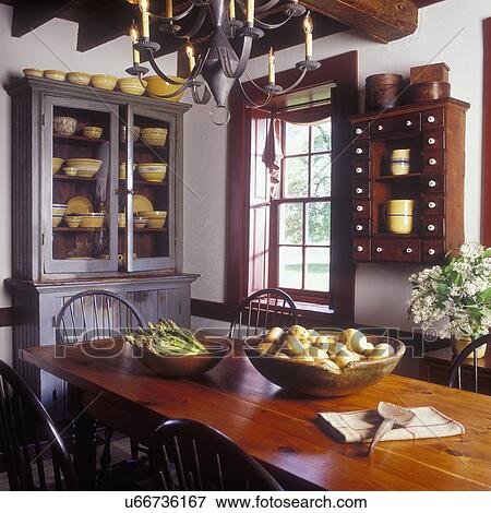 Stock Photo - DINING ROOMS - [Alternate view of dining area], angle shot, Plank table, white walls with cranberry trim, wooden bowls of asparagus and potatoes, hutch with collection of yellow ware, spice cabinet, swag, punched tin chandelier, lilacs, early American st. Fotosearch