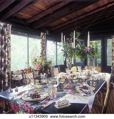Stock Photography - DINING ROOMS: Cottage look, vintage linens and tableware, enclosed porch, Hickory and cane twig chairs, summer set up on table, two bouquets of flowers, floral Barkcloth drapes, 1940's or 50's fabrics candle chandelier . Fotosearch