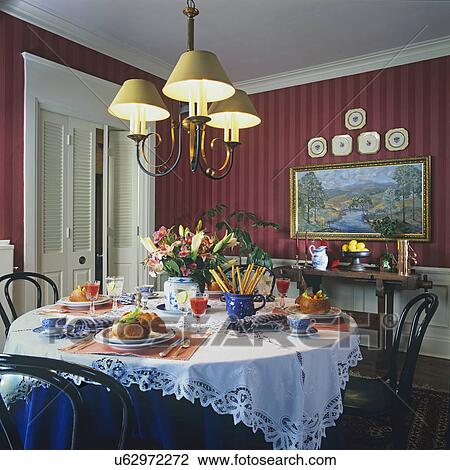 Stock Image - DINING ROOMS: Tone on tone red striped wallpaper, white crown molding and louvered folding doors, chandelier, round table in foreground, white cutwork lace tablecloth over sapphire blue cloth, table set with bread bowls,. Fotosearch