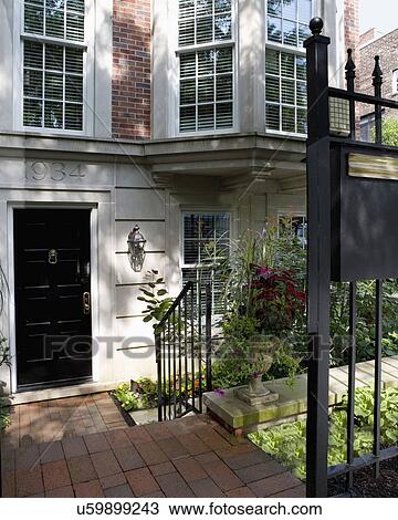 Stock Image - DOORS: EXTERIOR: city living, cut limestone with house numbers carved into stone, brick walkway, lanterns, step down from sidewalk, black railing, potted flowers, garden fence at sidewalk. Fotosearch