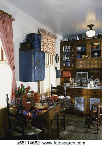 Stock Image - Eating area in kitchen with antique pantry cupboard, wooden cabinetry, tin ware, pierced tin panels, kitchen collectibles, hot cocoa and cookies on table, painted blue distressed wall cupboard. Fotosearch