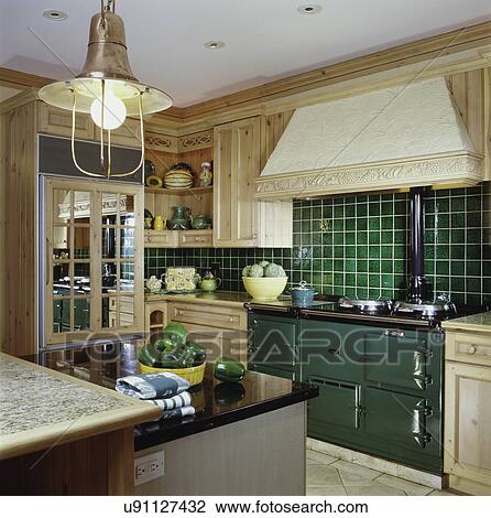 Stock Image - European style kitchen. Green ceramic tile, natural pine cabinets, mirror pane front on fridge, bowls on counters, large dark green Euro cooking range, stucco exhaust hood. Fotosearch
