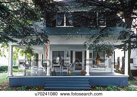 Stock Image - EXTERIOR: Pretty Summer cottage home with large front porch with columns. Porch all around. Dormer windows. American Flag, wicker carriage.. Fotosearch