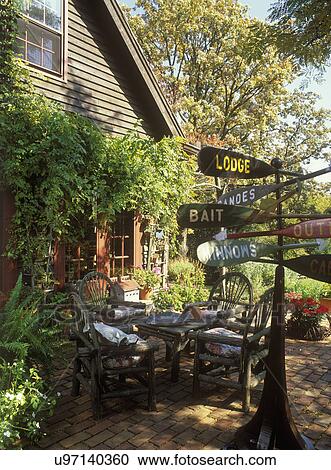 Stock Image - EXTERIORS - COUNTRY: Patio, twig furniture, signpost made of old oars, brick patio, climbing vines on side of country building, summer . Fotosearch