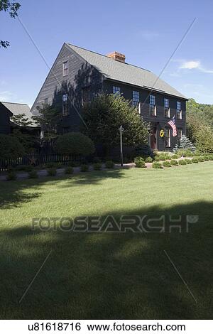 Stock Photograph - EXTERIORS: Dark brown salt box style home, sweeping lawns, gardens, American flag, vertical view with no garden in foreground. Fotosearch