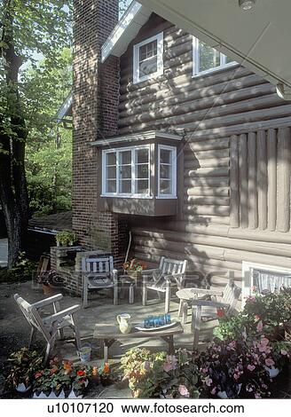 Stock Image - EXTERIORS - Log cabin style vacation home, white trim, brick chimney, Patio, wood chairs, Trees, potted flowering plants,. Fotosearch