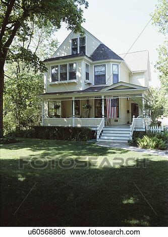 EXTERIORS: Three story remodeled/ restored Victorian, pale yellow clapboard, wraparound porch, American flag hangs from porch, hanging plants, wicker, shaded front lawn, no driveway showing, stone walk View Large Photo Image Stock Photograph - EXTERIORS: Three story remodeled/ restored Victorian, pale yellow clapboard, wraparound porch, American flag hangs from porch, hanging plants, wicker, shaded front lawn, no driveway showing, stone walk. Fotosearch
