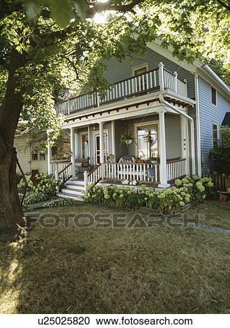 Stock Image - EXTERIORS: Two story single family farmhouse, two story porch, trimmed in white and burgundy, hydrangeas surround base, angle view. Fotosearch