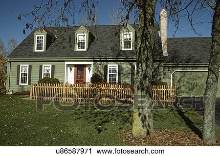 Stock Image - EXTERIORS: Two-story single family green Cape Cod with three dormers, natural picket fence, straight on view, attached garage to right and mature trees in right foreground. Fotosearch