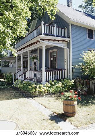 Stock Photo - EXTERIORS: Two story single family farmhouse, two story porch, trimmed in white and burgundy, hydrangeas surround base, barrel filled with geraniums, house next door can be seen. Fotosearch