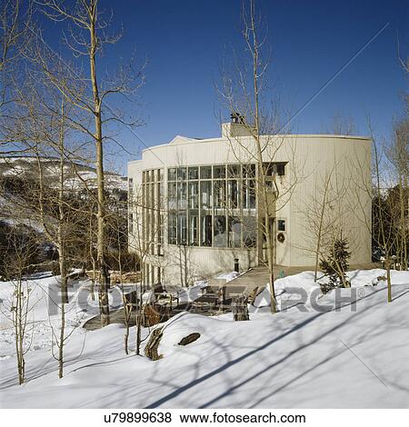 Stock Photo - EXTERIORS: white modern home nestled in mountains of Colorado, lots of windows, passive solar heating, winter time, snow, patio is cleared, outdoor seating. Fotosearch