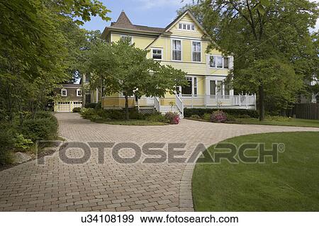 Stock Photo - EXTERIORS: Yellow clapboard Victorian with white trim, curved cobblestone driveway, three story home, detached garage, surrounded by mature trees, porch/deck in front, horizontal version. Fotosearch