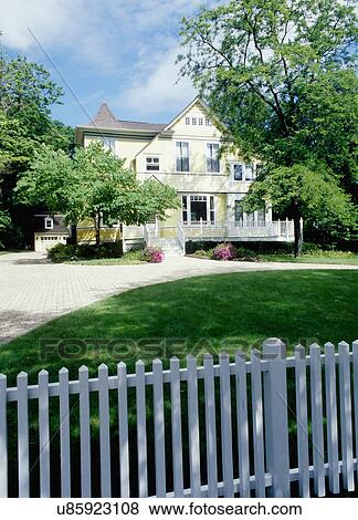 EXTERIORS: Yellow clapboard Victorian with white trim, curved cobblestone driveway, three story home, detached garage, surrounded by mature trees, porch/deck in front, vertical sunny white picket fence in foreground View Large Photo Image Stock Photo - EXTERIORS: Yellow clapboard Victorian with white trim, curved cobblestone driveway, three story home, detached garage, surrounded by mature trees, porch/deck in front, vertical sunny white picket fence in foreground. Fotosearch
