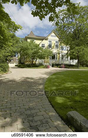 Stock Photograph - EXTERIORS: Yellow clapboard Victorian with white trim, curved cobblestone driveway, three story home, detached garage, surrounded by mature trees, porch/deck in front, vertical sunny. Fotosearch
