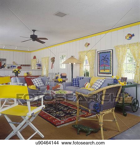 Picture - FAMILY ROOM - In pool house, white paneled walls, yellow trim, yellow tie back curtains, blue chambray sofas with multi colored pillows, pattern and sold, red blue yellow and white, directors chairs in yellow and white, summer beach feeling, ceiling fan,. Fotosearch
