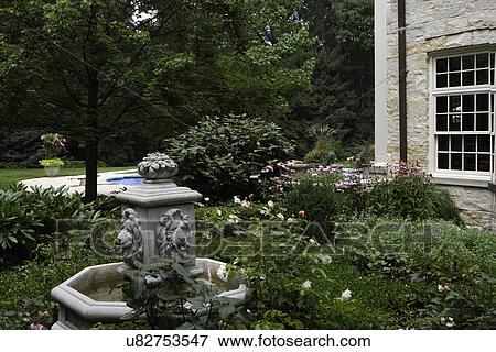 Stock Photo - GARDEN: foreground water fountain with lion heads as spouts, surrounded by boxwood and roses, coneflower, stone house to right, pool in background. Fotosearch