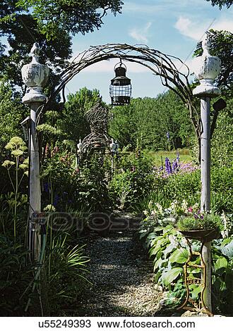 Stock Image - Gardens: Cedar twig and post arch defines opening to garden with hostas, hollyhocks, antique wooden finials, planter, delphiniums.. Fotosearch