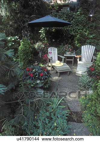 Picture - GARDENS: Close up of garden seating area, pink impatiens, green umbrella. Adirondack chairs, straw hat, pine trees, urn of impatiens flagstone path and patio.. Fotosearch