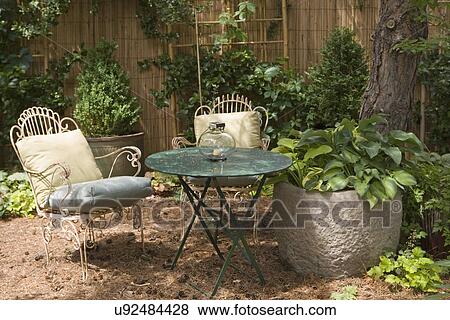 Stock Photo - GARDENS: pine needles create a patio base for a seating area, metal chairs and table, large concrete container filled with hosta, bamboo fence behind. Fotosearch