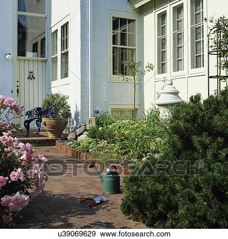 Stock Photo - GARDENS: Red brick walk along side of stucco house, border flower garden, watering can, pink rose bush, large urn planter.. Fotosearch