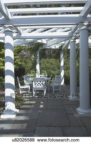 Stock Photo - GAZEBOS, PERGOLAS; Classic pergola, Chinese Chippendale style white table and chairs, slate patio, straight on view to eating area, three columns: one on left two to the right, two in the far distance behind patio set. vertical. Fotosearch