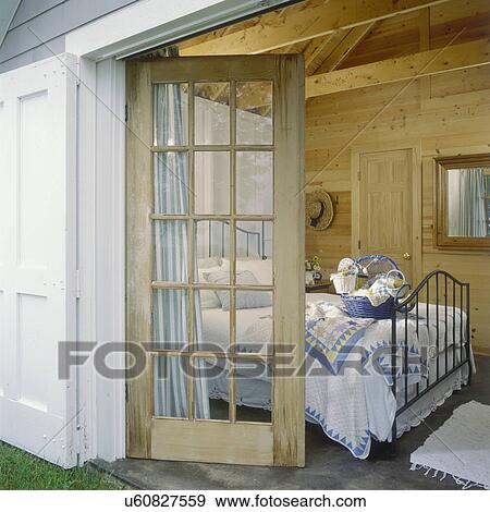 Stock Photo - GUEST BEDROOM - At lakefront cottage, kotty pine walls, exposed beams, cement floor, black iron bed with blue and white quilts, blue basket, mirror, striped curtains behind unpainted French door, looking from the outside in . Fotosearch