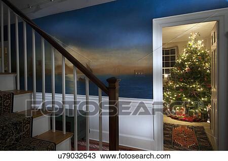Stock Image - Illuminated Christmas tree seen through doorway with painted wall and banister in house. Fotosearch