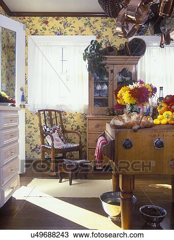 Stock Image - KITCHEN -Butcher block table with fall flowers, bread, vegetables, antique hutch with baskets, dishware, yellow floral wallpaper, copper pots hanging from rack, lace curtains, rocker with wooden footstool, wood floors, dog dishes, molding. Fotosearch