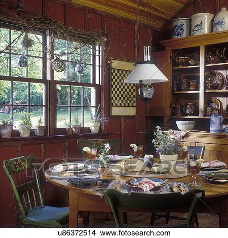 Kitchen Eating area, Bennington ware dishes. Rustic red walls, country styling, painted farm chairs, tole painted, game board, grapevine on window birdhouses hang from vine, plants in window, table set for lunch, hanging lantern View Large Photo Image Picture - Kitchen Eating area, Bennington ware dishes. Rustic red walls, country styling, painted farm chairs, tole painted, game board, grapevine on window birdhouses hang from vine, plants in window, table set for lunch, hanging lantern. Fotosearch