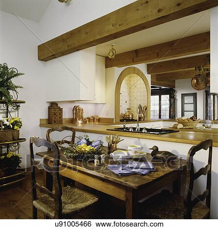 Stock Photograph - KITCHEN: Eating area in foreground, French provincial rustic chairs and farm table, open to kitchen with light stained pine wood counters, arched exhaust hood over stove area, contemporary home with rustic country accents. Fotosearch
