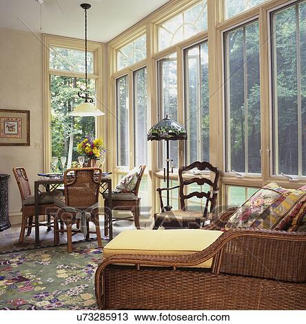 Stock Image - KITCHEN EATING AREA: Open family room area with floor to ceiling windows, four rattan chairs sit around a square table, chaise lounge in foreground, French provincial chair stained glass lamp. Fotosearch