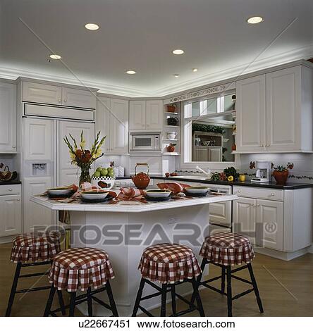 Stock Image - KITCHEN: gingham check red and white fabric on four stools in foreground, kitchen eating area, recessed indirect lighting above white cabinets, pass through over sink area. Fotosearch