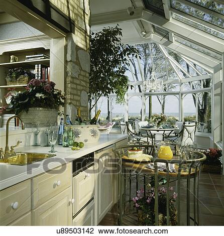 Stock Image - KITCHEN: Serving area, bras bar sink, serving cart with wine, champagne and cheese, tile floor, brick and stone wall, looking onto eating area in solarium, table with place settings. Fotosearch