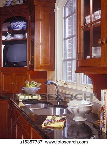 Stock Photo - KITCHEN SINK AREA - View of sink counter area with granite counter top and cherry cabinetry with small TV behind doors.. Fotosearch