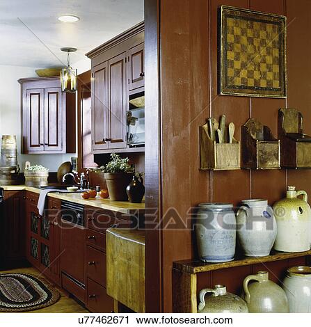 Stock Image - KITCHEN: View into kitchen, red painted pine cabinets, maple counter tops, 1800's salt glaze crocks on shelves in foreground right, wooden salt boxes with wooden spoons, game board, contemporary period style country. Fotosearch