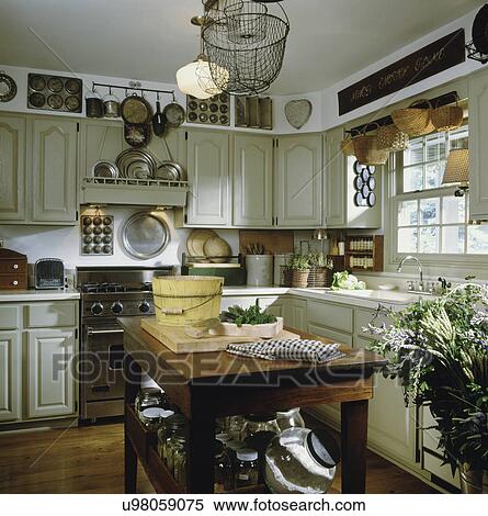 Stock Photography - Kitchen with counter top, sink, cabinets, stove, island areas, herbs on counter, pewter & tin cooking utensils on display, hanging light, and wood floor. Fotosearch