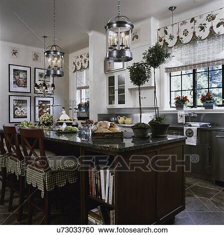 Stock Image - Kitchen with cream walls, vintage magazine advertising framed,, topiaries, window valance, tall bar chairs with checkered fabric skirts and walnut stained island, stainless steel counters and backsplash. Fotosearch
